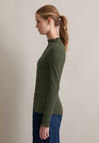 Langemouwen top in olijfgroen met een hoge, gerimpelde halslijn en geribbelde textuur. De zoom is licht gegolfd, gecombineerd met een blauwe jeans.