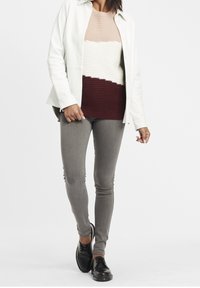 Pull en maille à rayures beige, blanc et bordeaux, superposé sous une chemise boutonnée vert clair. Pantalon gris slim et chaussures noires.