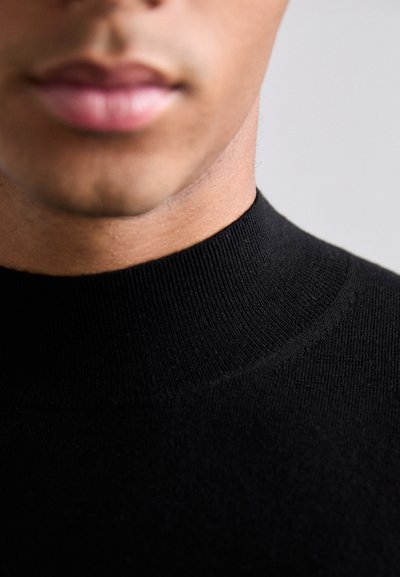 Schwarzer gerippter Rollkragensweater mit glatter Textur, der ein figurbetontes Design und einen hohen Kragen aufweist, wodurch der Kragen- und obere Brustbereich betont wird.