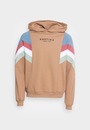 Sweat à capuche beige avec des rayures chevrons bleues, rouges, blanches et vertes sur les manches et le texte "KAOTIKO Since 1999" sur le devant de la poitrine.
