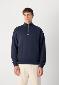 Sudadera con cremallera en color navy, con cuello alto, puños y dobladillo acanalados. Presenta un pequeño logo azul en el pecho como elemento de marca. Textura suave.