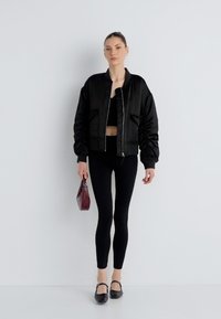 Μαύρο μπουφάν bomber με ριμπ μανσέτες, συνδυασμένο με μαύρο crop top και κολάν. Το μοντέλο κρατά μια μικρή μπορντό τσάντα χειρός. Μαύρα επίπεδα παπούτσια.