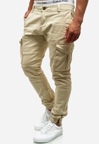 Pantalons cargo beiges avec poches latérales, poignets élastiques et une coupe ajustée. Portés avec des baskets blanches et une ceinture de marque visible.