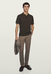 Polo di colore marrone scuro con maniche corte, abbinata a pantaloni marrone chiaro e scarpe marroni. Tiene in mano un maglione lavorato a maglia grigio. Design semplice.