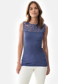 Haut sans manches bleu avec un panneau en dentelle florale au niveau du col, texture côtelée et design ajusté. Porté avec un pantalon blanc.