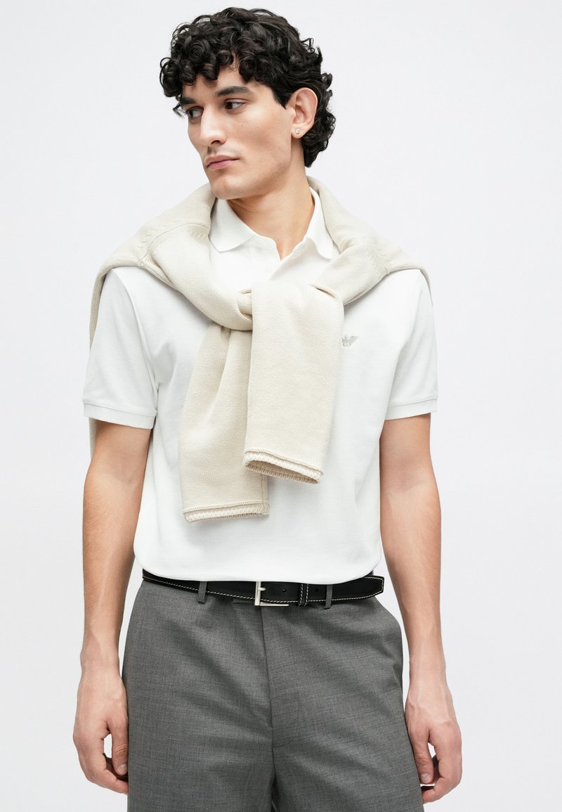 Weißes Poloshirt, leichter hellbeiger Strickpullover lässig über die Schultern gelegt, graue maßgeschneiderte Hose, schwarzer Gürtel mit Metallschnalle, glatte Texturen.