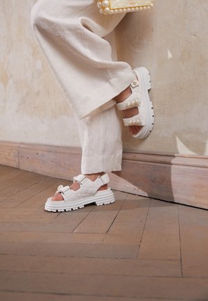 Persona vistiendo pantalones blancos de pierna ancha y sandalias blancas gruesas con adornos de perlas, de pie sobre un suelo de madera frente a una pared pálida.