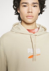 Beige hettegensere laget av myk stoff, med kengurulomme, snorhette og brodert "PUMA"-logo i hvite og oransje akenter.