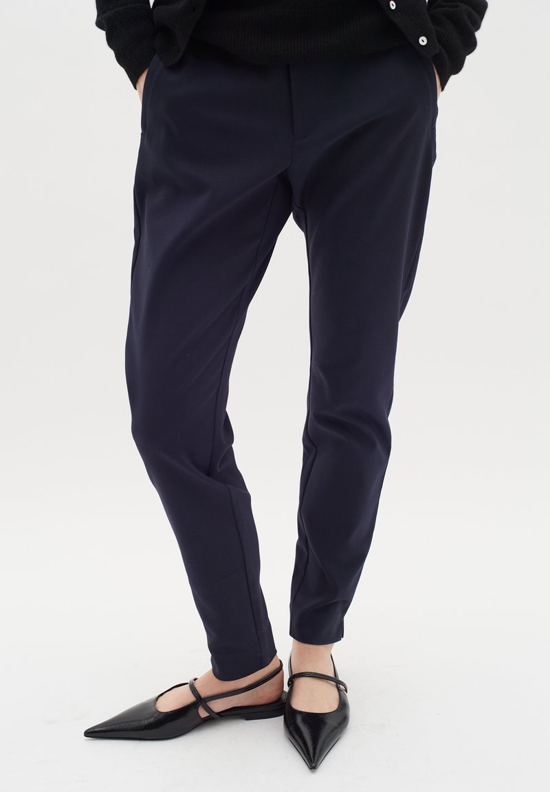 Pantalon marine avec une coupe ajustée, texture lisse et jambes fuselées, associé à des chaussures plates noires avec des orteils pointus et des brides de cheville.
