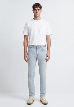 Young man standing facing forward wearing a white short-sleeve T-shirt, light blue jeans, and beige sneakers against plain background.