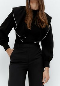 Černý top s dlouhým rukávem, který má volánkový design a kamínky po okrajích, ve spojení s oblekovými černými kalhotami.