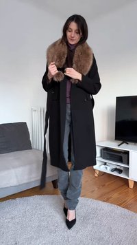 Black long coat with a large brown faux fur collar, a double-breasted design, belted waist, paired with gray jeans and black pointed shoes.