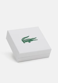 Boîte cadeau blanche avec une finition texturée, arborant un logo crocodile vert sur le couvercle. La boîte est carrée et légèrement relevée à un coin.
