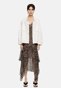 Veste en denim blanc avec quatre poches sur une robe à imprimé léopard marron présentant un ourlet asymétrique. Sandales noires avec des accents décoratifs.