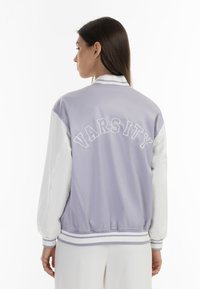 Lila varsityjack met witte mouwen, met geborduurde "VARSITY" tekst op de achterkant, geribbelde manchetten en een gestreepte zoom. Gladde textuur.