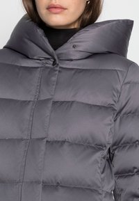 Chaqueta acolchada gris con un cuello alto y sobredimensionado, con costuras acolchadas y cierre de presión, fabricada con un material liso y ligero.