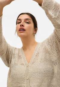 Cardigan in maglina beige chiaro con scollo a V, dettagli in paillettes e un motivo di trama aperta. Presenta chiusura con bottoni e maniche ampie.