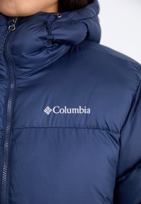Tamno plava puferska jakna s teksturiranim, quiltanim završetkom. Sadrži visoki ovratnik, prednji zatvarač i bijeli Columbia logo na prsima.