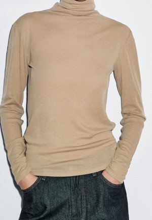 Personne portant un pull à col roulé beige à manches longues et un jean en denim foncé, les mains dans les poches, sur un fond uni.