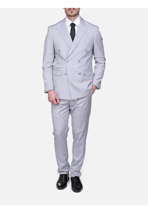 Homme portant un costume croisé gris clair avec une chemise blanche, une cravate noire et des chaussures habillées noires, debout devant un fond uni.