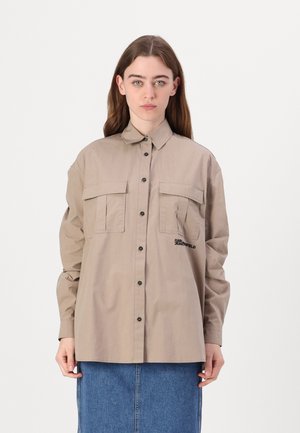 Beige button-up shirt with two chest pockets, black buttons, and embroidered text on the left side. Cotton fabric with a smooth texture.