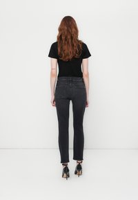 Femme aux longs cheveux roux portant un t-shirt noir, un jean gris foncé et des chaussures à talons noirs, se tenant face à un mur blanc.