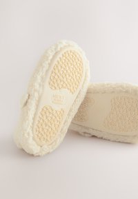 Chaussons en peluche blanche avec une semelle en caoutchouc texturée et une tige douce et duveteuse. Détails de l'étiquette visibles sur les semelles intérieures.