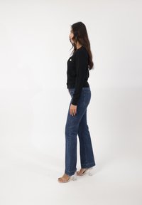 Cárdigan negro de canalé con detalle de botones, combinado con jeans de mezclilla azul de pierna ancha y sandalias de tacón transparente. Texturas suaves, ajuste casual.