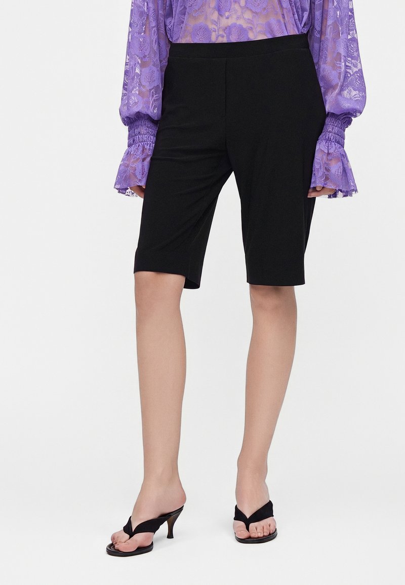 Personne portant un short noir au genou, un chemisier en dentelle violette transparente à manches longues, et des sandales noires à talons, debout sur fond blanc.