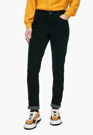 Person iført sorte opkørte bukser, hvide og orange sneakers og en gul langærmet skjorte med den ene hånd i lommen og den anden afslappet.