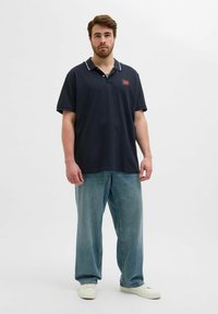 Ein marineblaues Poloshirt mit weißem Kragenbesatz und Logo-Patch, kombiniert mit lockeren hellblauen Jeans und weißen Sneakers, vor einem schlichten Hintergrund stehend.
