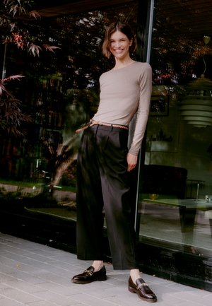 Beige Langarmoberteil, schwarze weite Hose, brauner geflochtener Gürtel und dunkle Slipper. Außeneinstellung mit gläsernem Hintergrund und Grünanlagen.