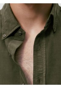 Gros plan de la partie supérieure de la poitrine et du cou d'une personne portant une chemise olive ouverte, montrant des poils légers sur la poitrine.