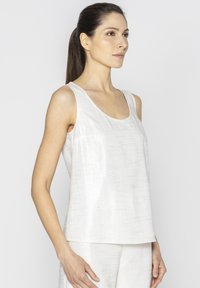 Biała, bezrękawnikowa koszulka wykonana z teksturowanej tkaniny, z szerokim dekoltem i luźnym krojem. Prezentowana na modelce w profilu.