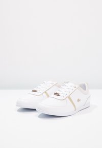 Bílé sportovní boty se zlatými akcenty, vyrobené z hladkého a perforovaného syntetického materiálu; mají šněrování a logo na boku.