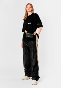 Camiseta negra de gran tamaño con logo blanco, combinada con vaqueros holgados negros con rayas laterales y un bolso cinturón de color beige. Zapatos blancos gruesos.