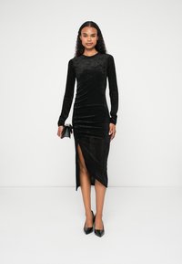 Μαύρο βελούδινο φόρεμα με μακρύ μανίκι, σχισμή στο πλάι, εφαρμοστό σχέδιο και μαλακή υφή. Το μοντέλο κρατά μια μικρή, μαύρη τσάντα χεριού. Μαύρες μυτερές γόβες.