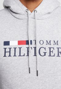 Sudadera gris con un bolsillo tipo canguro. Presenta un gran logo azul "HILFIGER" y una franja horizontal roja, blanca y azul encima de él.