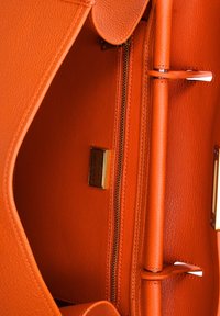 Interieur van een oranje leren handtas met een textuurafwerking, voorzien van een ritsvak en een metallic merklabel. Strakke lijnen en een minimalistisch ontwerp.