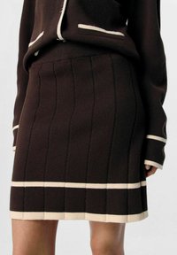Jupe en tricot marron avec une texture à côtes verticales et des rayures horizontales beiges au niveau de l'ourlet et de la taille. Fini avec un passepoil beige contrastant.