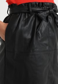 Jupe en cuir noir avec une taille froncée et une ceinture nouée, poches latérales et texture lisse, mettant en valeur des détails de couture.