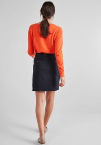 Pull orange en tricot associé à une jupe ajustée bleu marine, avec des poches à l'arrière et une texture lisse. Porté avec des chaussures plates à motifs.
