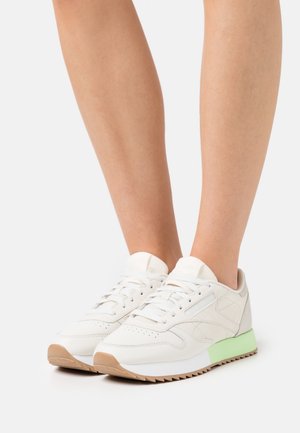 Persona con zapatillas bajas blancas con suela de color verde claro y caramelo, de pie sobre un fondo blanco.