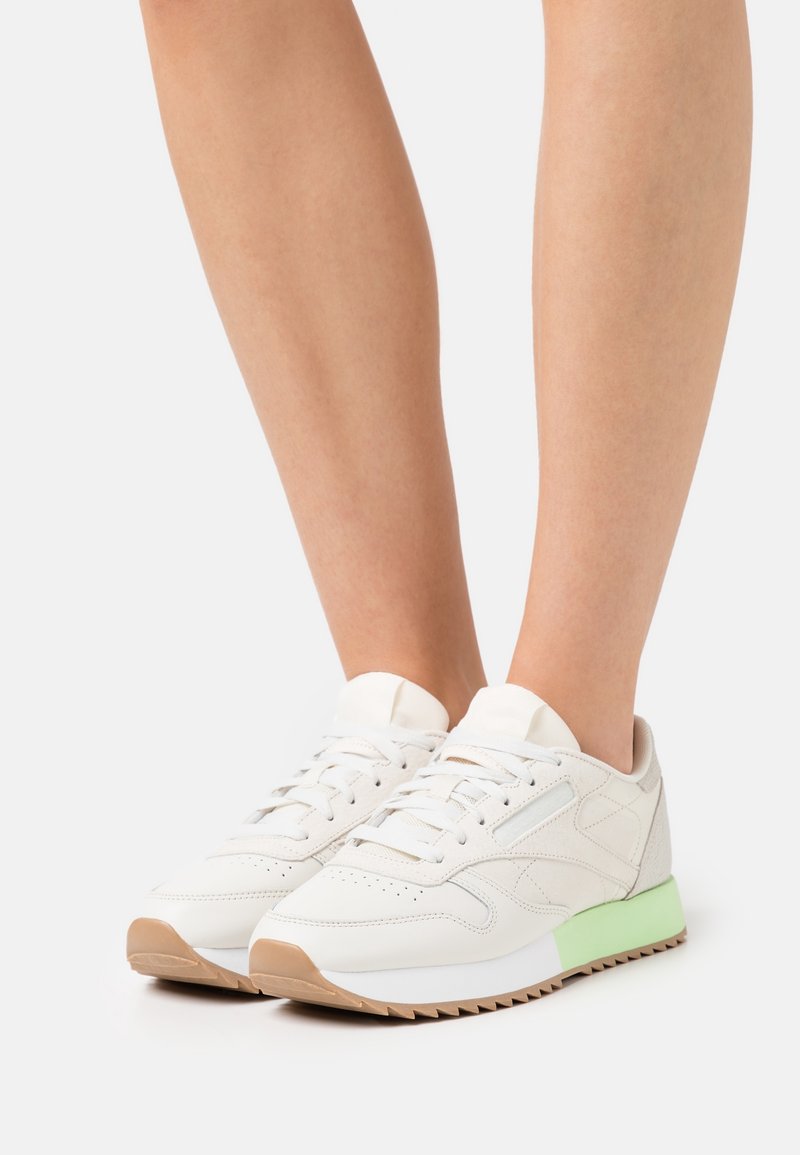 Personne portant des baskets basses blanches avec une semelle vert clair et gomme, debout sur un fond blanc.