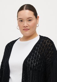 Black open-knit cardigan with a textured, diamond pattern. Lightweight fabric, featuring wide sleeves and a relaxed fit over a white top.