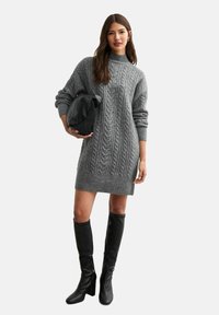 Abito in maglia grigia con motivo a treccia, collo alto e polsini a costine. Indossato con stivali neri fino al ginocchio e una borsa nera.