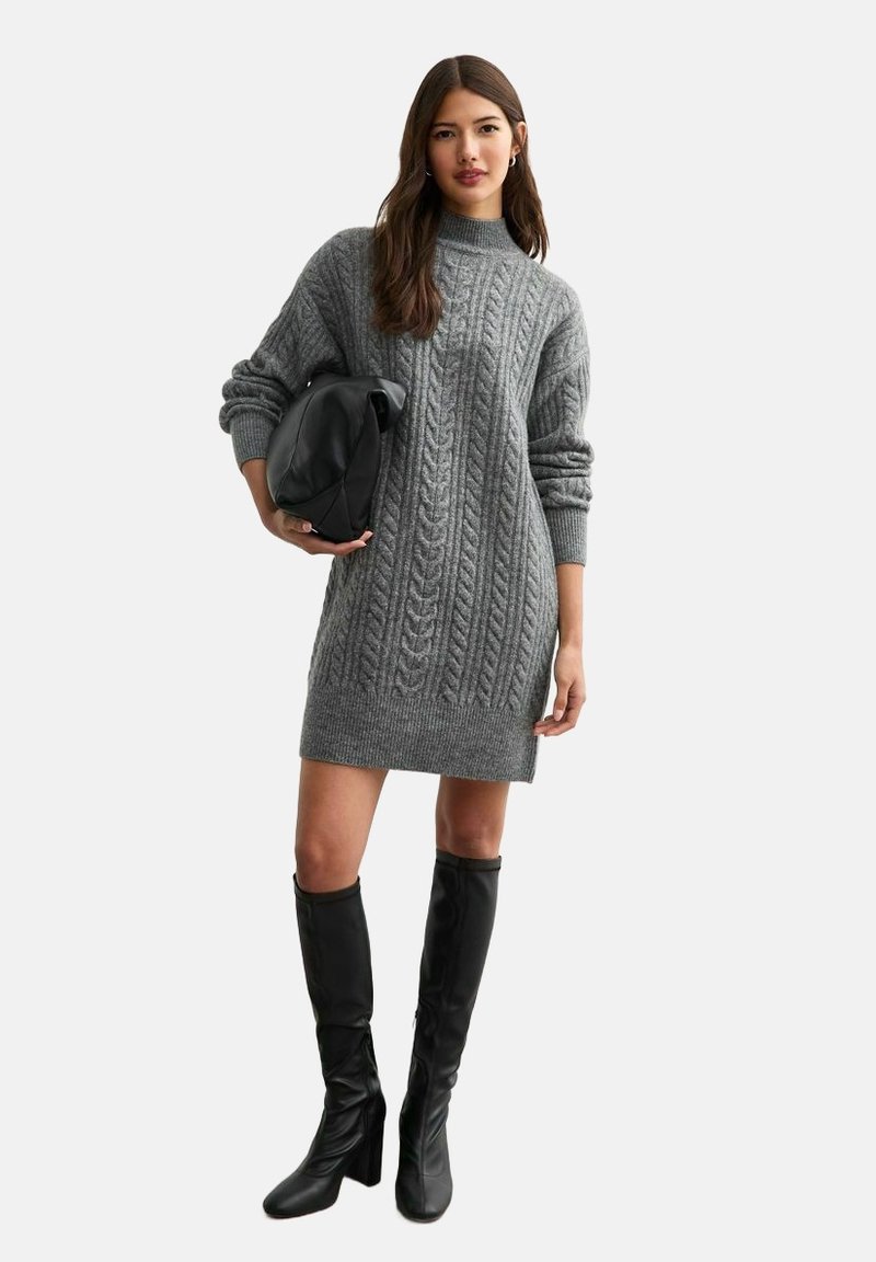 Abito in maglia grigia con motivo a treccia, collo alto e polsini a costine. Indossato con stivali neri fino al ginocchio e una borsa nera.