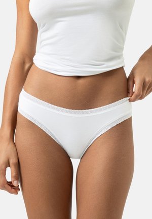 Nainen, joka pukeutuu valkoiseen pitsireunuksella koristeltuun puuvillaisiin alusvaatteisiin ja valkoiseen toppiin, säätää vyötärönauhaa yhdellä kädellä yksiväristä taustaa vasten.