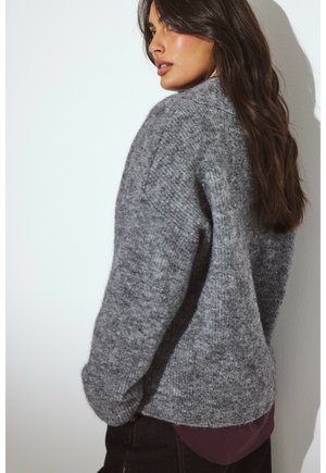 Frau mit langen, dunklen Haaren, die einen lockeren grauen Strickpullover über einem dunkelmaroonen Shirt trägt und seitlich gegen eine helle Wand steht.