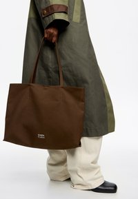 Ruda drobės pintinė su dviem rankenomis, pasižyminti minimalizmo dizainu ir logotipu, spausdintu apačioje. Dėvima su žaliu paltu ir smėlio spalvos kelnėmis.